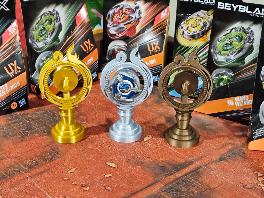 Three Beyblade trophies in gold, silver, and bronze displayed against a colorful background of Beyblade X packaging.